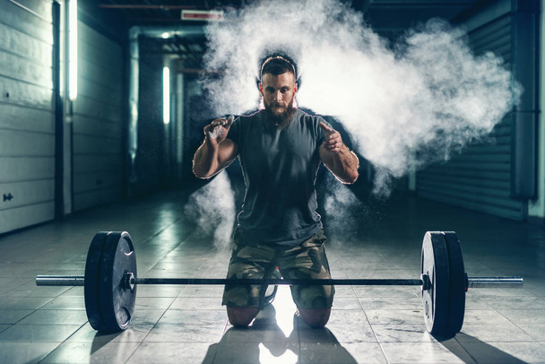 Strong muscular Caucasian bodybuilder clapping hands and crouching in hall. In front of him barbell, chalk all around. Back light, night workout concept. - Photo, Image