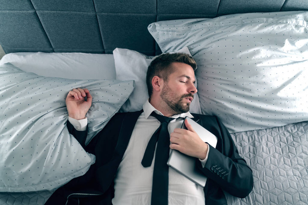 Przepracowany biznesmen w garnitur, spanie i leżąc na łóżku i trzymając tablet na jego klatce piersiowej. - Zdjęcie, obraz