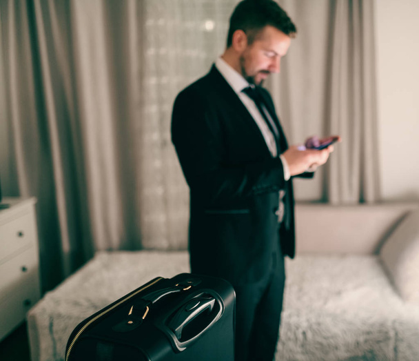 Uomo d'affari che utilizza lo smartphone mentre si trova in camera da letto e si prepara per il viaggio. Focus selettivo sulla valigia
. - Foto, immagini