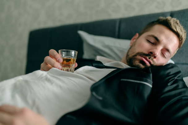 Sleeping businessman sleeping in the bed in hotel room and holding alcohol shot on the chest. Alcohol abuse concept. - Foto, immagini