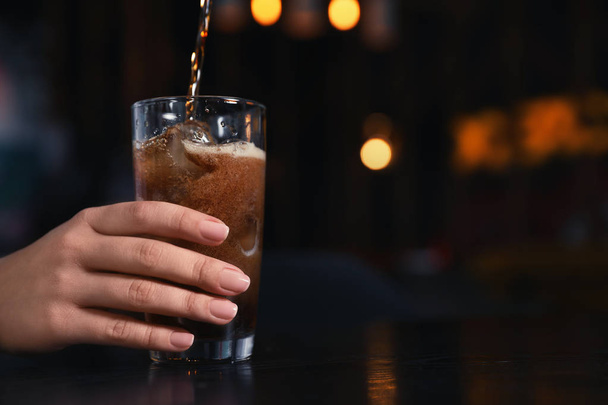 Woman holding glass with ice cubes while pouring cola at table indoors, closeup. Space for text - Φωτογραφία, εικόνα