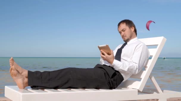 Νεαρός επιχειρηματίας εργάζεται στην παραλία. Ανδρική επαγγελματική κάθεται σε άνετη ξαπλώστρα - Πλάνα, βίντεο