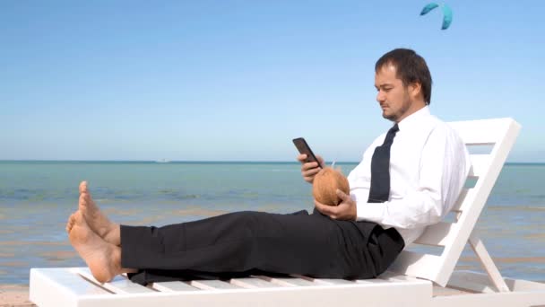 Geschäftsmann sitzt im Anzug mit einem Cocktail auf einer Liege vor dem Hintergrund eines schönen Meeres - Filmmaterial, Video