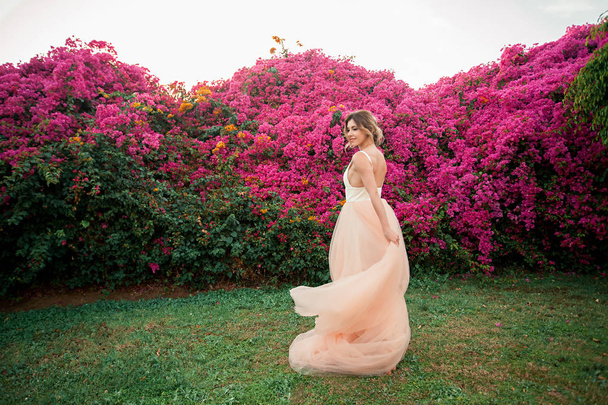 Beautiful bride in a wedding dress with a long veil laughs against a background of pink flowers - Foto, Imagen