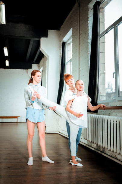 Long-haired blond teenager in white sport clothing doing legs stretching near the ballet bar - Foto, afbeelding