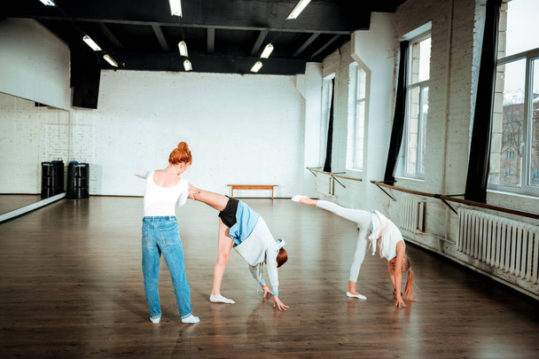 Red-haired dance teacher in blue jeans correcting leg position of her student - Фото, зображення