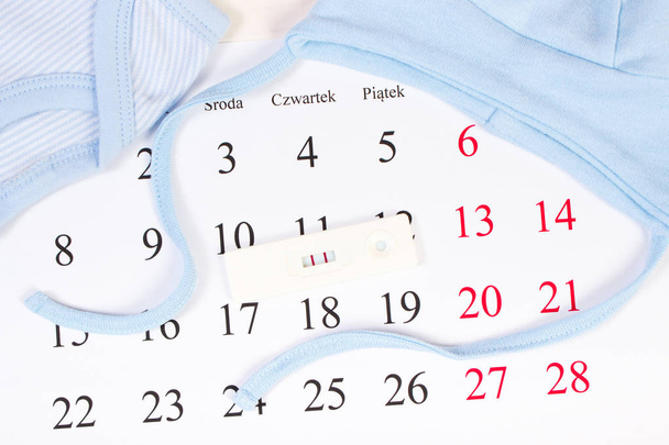 Zwangerschapstest met positief resultaat en kleding voor pasgeboren op kalender met Pools inscriptie, familie uit te breiden en verwacht voor baby concept - Foto, afbeelding