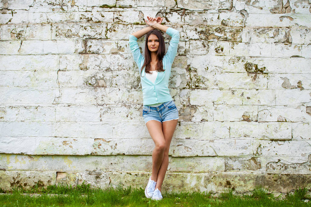 Young beautiful brunette woman in short blue jeans shorts posing against a stone wall background - Valokuva, kuva