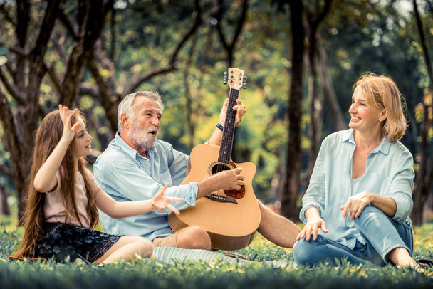 Ευτυχισμένη οικογένεια παίζει κιθάρα και να τραγουδήσει μαζί ενώ κάθεται στο πάρκο το καλοκαίρι. Έννοια της οικογένειας συγκόλληση από μουσική. - Φωτογραφία, εικόνα
