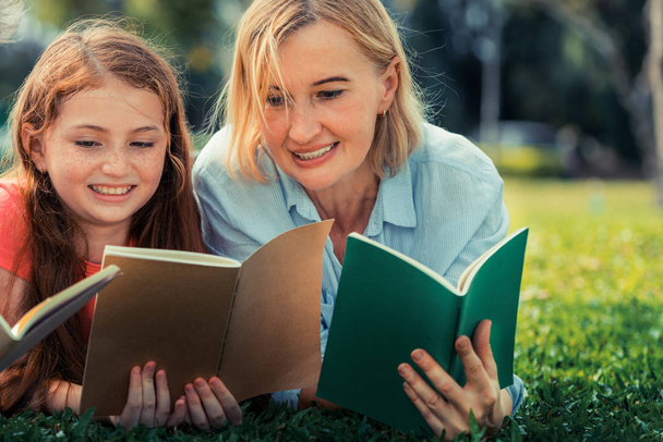 Happy family read books together and lying on green grass in public park. Little girl kid learning with mother in outdoors garden. Education and family lifestyle. - 写真・画像