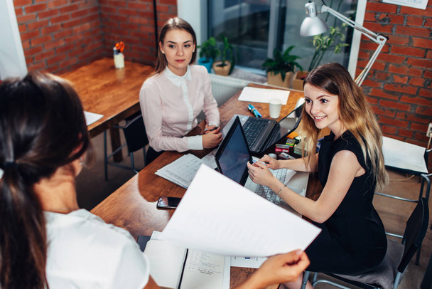 Команда деловых женщин, работающих с документами, используя ноутбуки, сидящие за столом в офисе
 - Фото, изображение