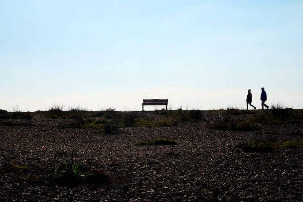 im Vordergrund ist ein Kieselstrand am Ende des Strandes sind zwei nicht wiedererkennbare Menschen, die als Paar zusammen gehen, sie werden von der Sonne und dem Himmel dahinter einsam gemacht, sie gehen auf eine Parkbank zu, die ebenfalls einsam ist, Shoreham, East susse - Foto, Bild