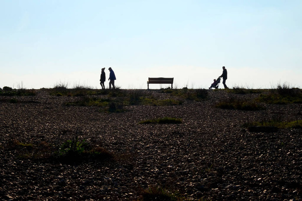 手前では、ビーチの端に小石のビーチは歩いて 3 つの認識できない人々、左側真ん中にカップルは公園のベンチ、右側の puch 椅子で子供と父、彼らは太陽の下ですべての silouetted には、 - 写真・画像