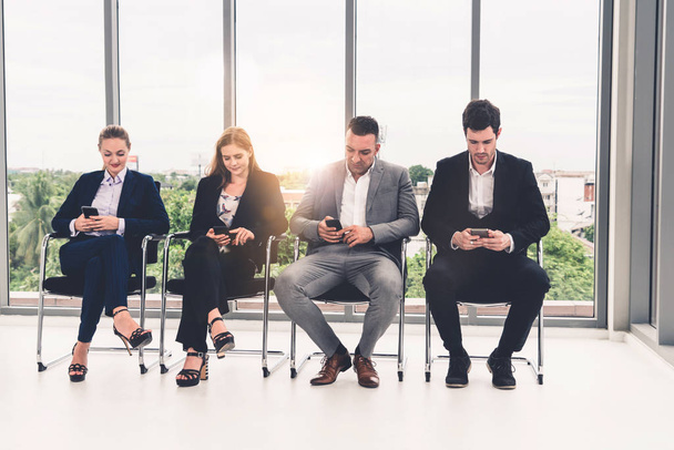 Empresárias e empresários usando telefone celular enquanto esperam em cadeiras no escritório para entrevista de emprego. Conceito de negócios corporativos e recursos humanos
. - Foto, Imagem