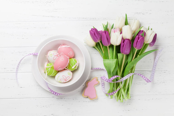 Pasen-wenskaart met tulp bloemen boeket, paaseieren en konijnen cookie. Bovenaanzicht over witte houten tafel - Foto, afbeelding