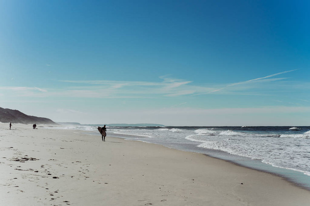 man surfer on the beach with surfboard, walks along the ocean shore, Nazar, Portugal. - 写真・画像