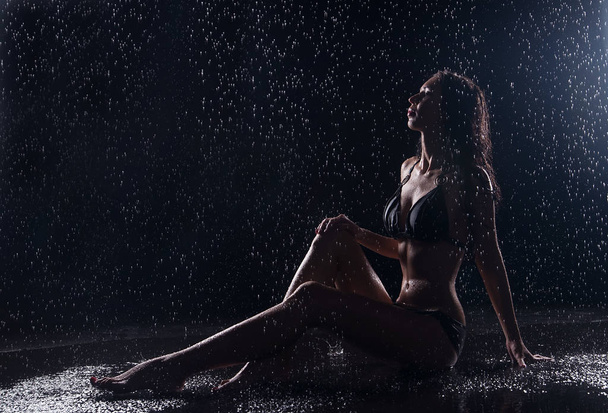 Young woman in black swimsuit sitting on wet black background  - Photo, Image