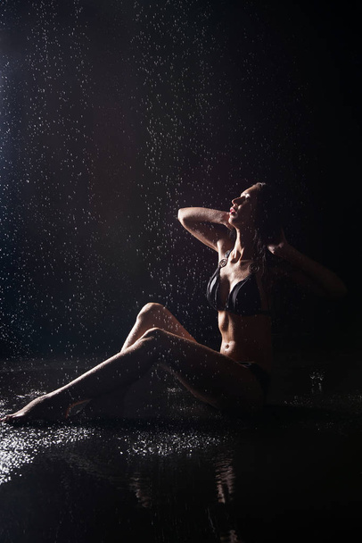 Молодая женщина в черном купальнике сидит на мокром черном фоне
  - Фото, изображение