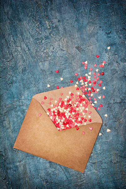 Craft paper envelope for romantic love letter with pink sweet sugar sprinkles or confetti in heart shape on blue concrete background with copy space - Foto, afbeelding