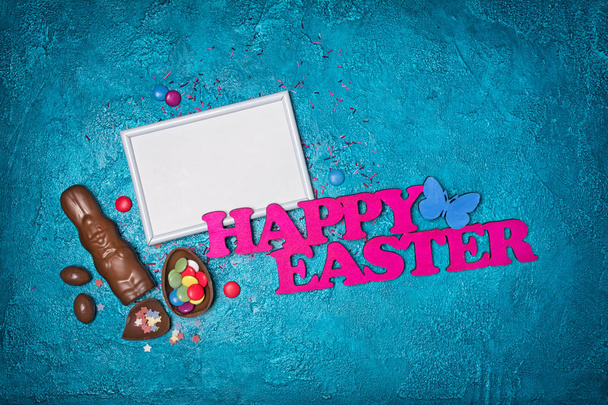 Top view on mockup of blank white frame with pink text of happy easter and traditional sweets as chocolate eggs and colorful sugar sprinkles on turquoise concrete background - Foto, Imagem
