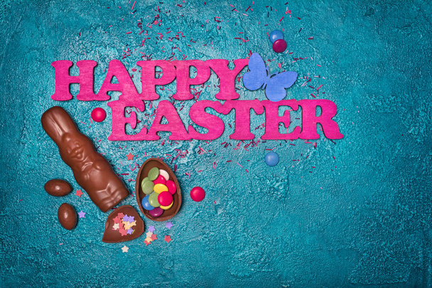 Composition of pink text of happy easter with chocolate traditional eggs and bunnies with bright colorful dragee and sugar sprinkles on turquoise concrete background - Photo, image