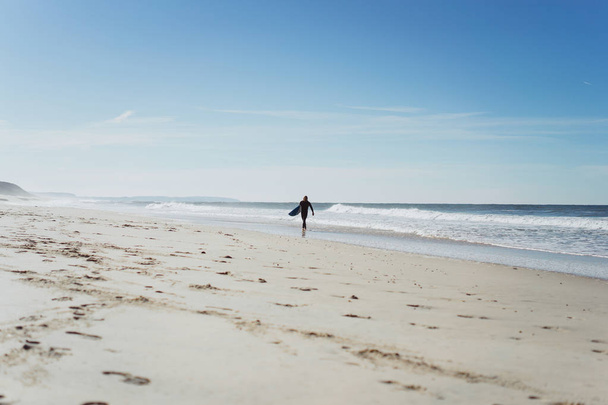 Hombre con tabla de surf en la orilla del océano. Surfista con traje de neopreno. Nazare, Portugal
. - Foto, Imagen