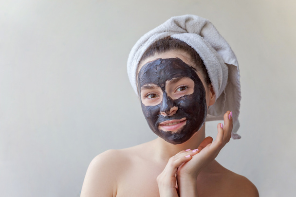 Portret van de schoonheid van een lachende brunette vrouw in een handdoek op het hoofd zwart voedende masker aanbrengen op gezicht op witte achtergrond geïsoleerd. Huidverzorging reinigende spa relax cosmetica concept - Foto, afbeelding