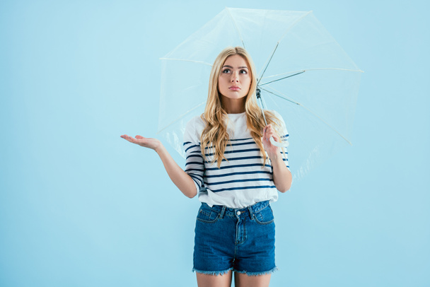 Molesto chica rubia de pie bajo el paraguas sobre fondo azul
 - Foto, Imagen