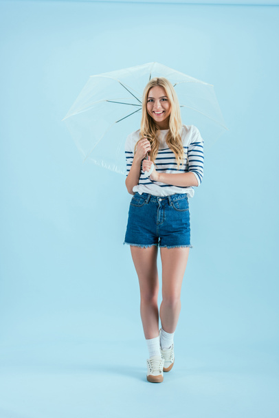 Joyful blonde girl holding umbrella on blue background - Fotó, kép