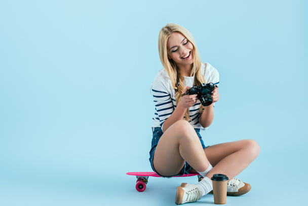 Smiling girl sitting on pink longboard and holding camera on blue background - Foto, immagini