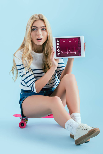 Shocked girl holding digital tablet with fitness app on screen and sitting on pink longboard on blue background - Fotografie, Obrázek