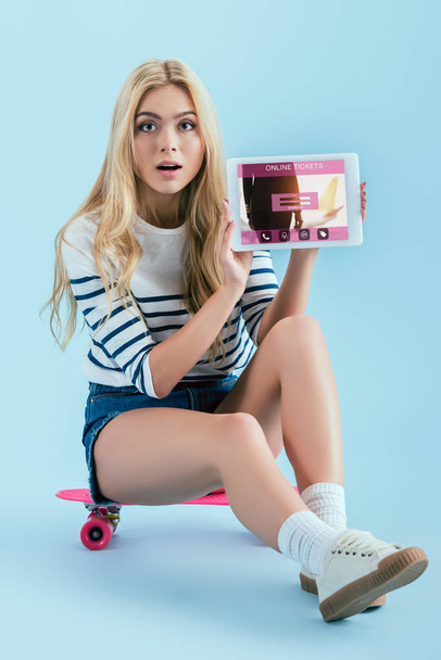Surpreendida menina de cabelos longos mostrando tablet digital com aplicativo de bilhetes on-line na tela e sentado em longboard no fundo azul
 - Foto, Imagem