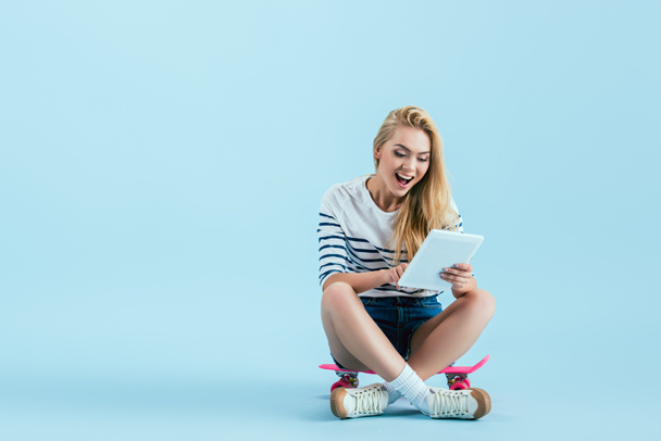 Izgatott lány segítségével digitális tabletta, ülve longboard a kék háttér - Fotó, kép