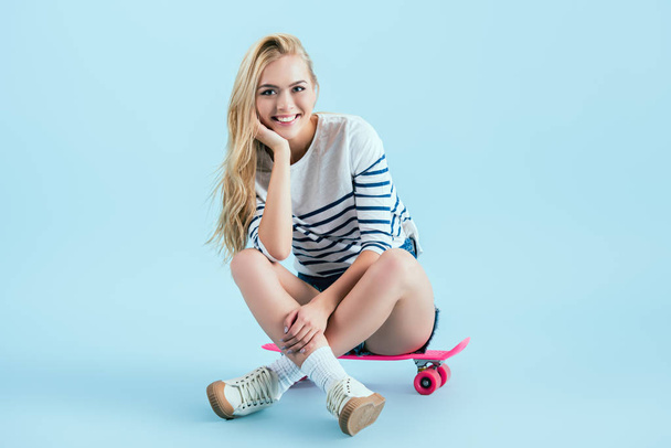 Smiling girl sitting on longboard with legs crossed on blue background - Foto, afbeelding