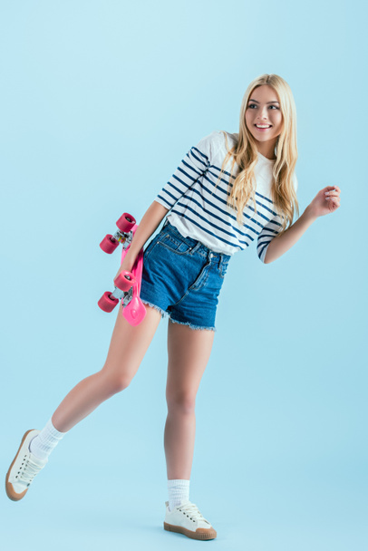 Smiling girl with longboard standing on one leg isolated on blue - Фото, зображення