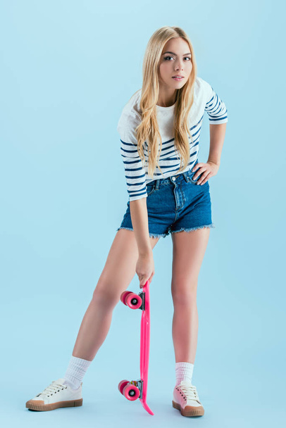 Charming blonde girl posing with pink longboard isolated on blue - Fotó, kép