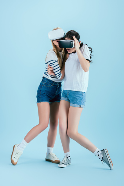 Studio shot of surprised girls in VR headsets embracing on blue background - Фото, зображення