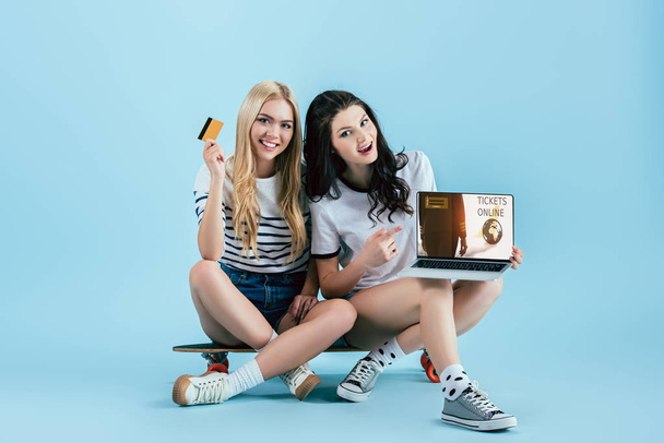 Κορίτσια για longboard κρατώντας laptop με εισιτήρια online ιστοσελίδα στην οθόνη και την πιστωτική κάρτα σε μπλε φόντο - Φωτογραφία, εικόνα