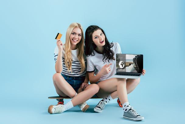 Smiling girls holding laptop with booking website on screen and credit card while sitting on longboard on blue background - Фото, изображение