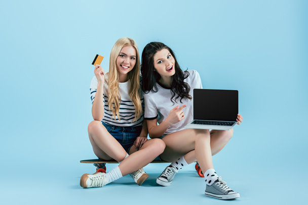 Mooie meisjes zitten op longboard met credit card en laptop met leeg scherm op blauwe achtergrond - Foto, afbeelding