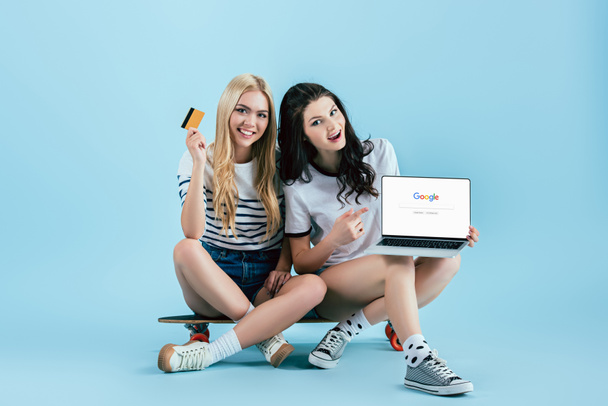 Longboard üzerinde oturan ve laptop ile google web sitesinde ekran ve kredi kartı mavi arka plan üzerinde tutarak çarpıcı kız - Fotoğraf, Görsel