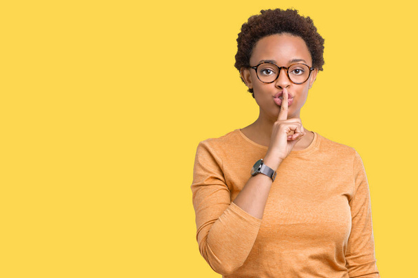junge schöne afrikanisch-amerikanische Frau trägt eine Brille über isoliertem Hintergrund und bittet um Ruhe mit dem Finger auf den Lippen. Schweigen und geheimes Konzept. - Foto, Bild