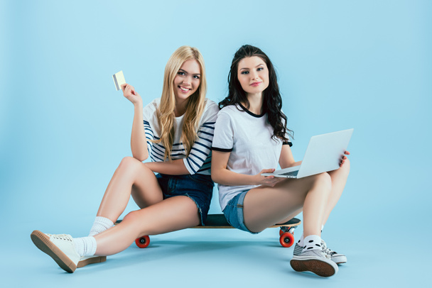 Jolies filles avec ordinateur portable et carte de crédit assis sur longboard sur fond bleu
 - Photo, image