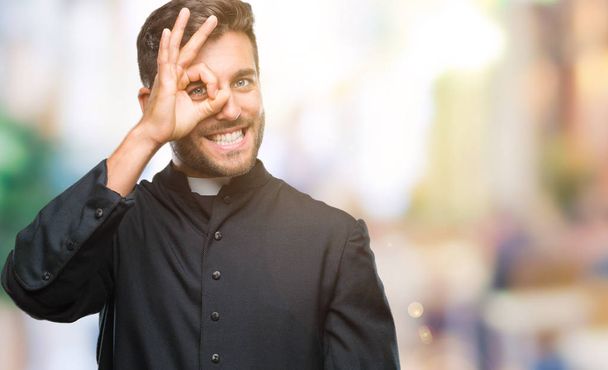 Mladý kněz křesťanské muž nad izolované pozadí dělá ok gesto rukou, s úsměvem, oko dívá skrze prsty s šťastný obličej. - Fotografie, Obrázek