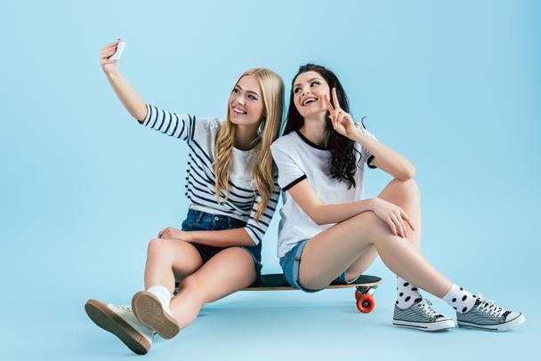 Stylish girls sitting on longboard and taking selfie with peace sign on blue background - Photo, Image