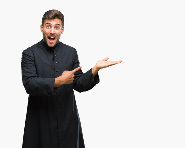 Молодой католический священник-христианин на изолированном фоне удивился и улыбнулся в камеру, показывая рукой и указывая пальцем
. - Фото, изображение