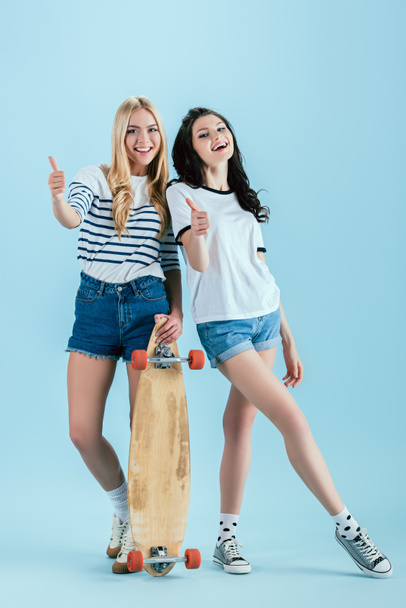 Stylish girls with wooden longboard posing with thumbs up on blue background - Photo, Image