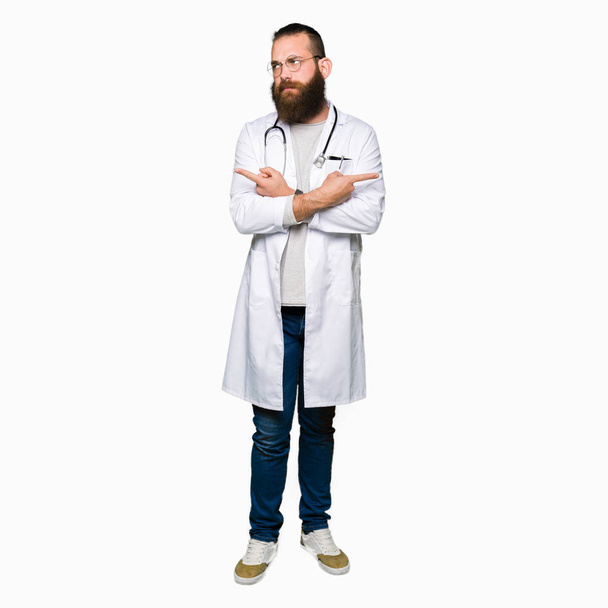 Joven médico rubio con barba usando abrigo médico Señalando a ambos lados con los dedos, dirección diferente en desacuerdo
 - Foto, imagen