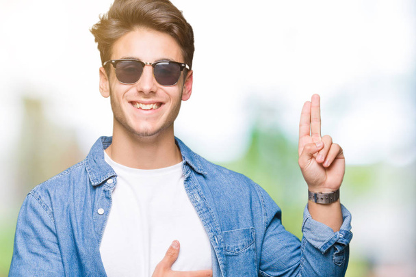Mladý pohledný muž nosí sluneční brýle přes izolované na pozadí Swearing s rukou na hrudi a prstech, takže slib slib věrnosti - Fotografie, Obrázek