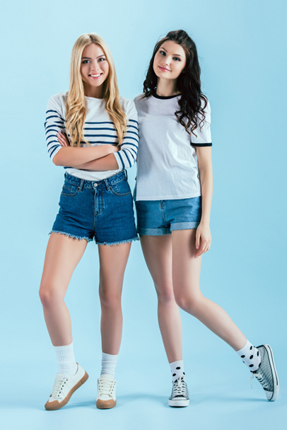 fröhliche Mädchen in Jeanshosen blicken in die Kamera auf blauem Hintergrund - Foto, Bild